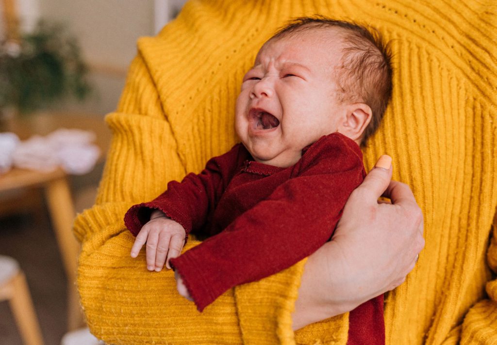 A crying baby wearing a red outfit is held by someone wearing a gold sweater.