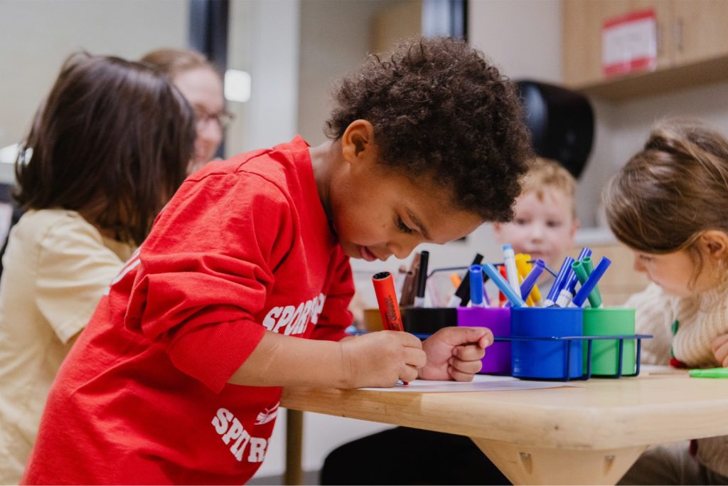 A young boy wearing a red shirt writes with a red marker at a table with other children at the Child Development Lab.