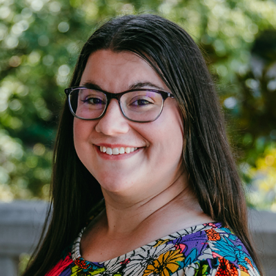 A woman with glasses and long brown hai wearing a multicolored shirt.