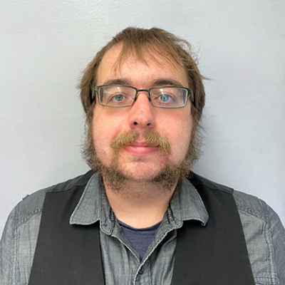 A man with brown hair with a mustache and beard wearing a grey shirt and a black vest.