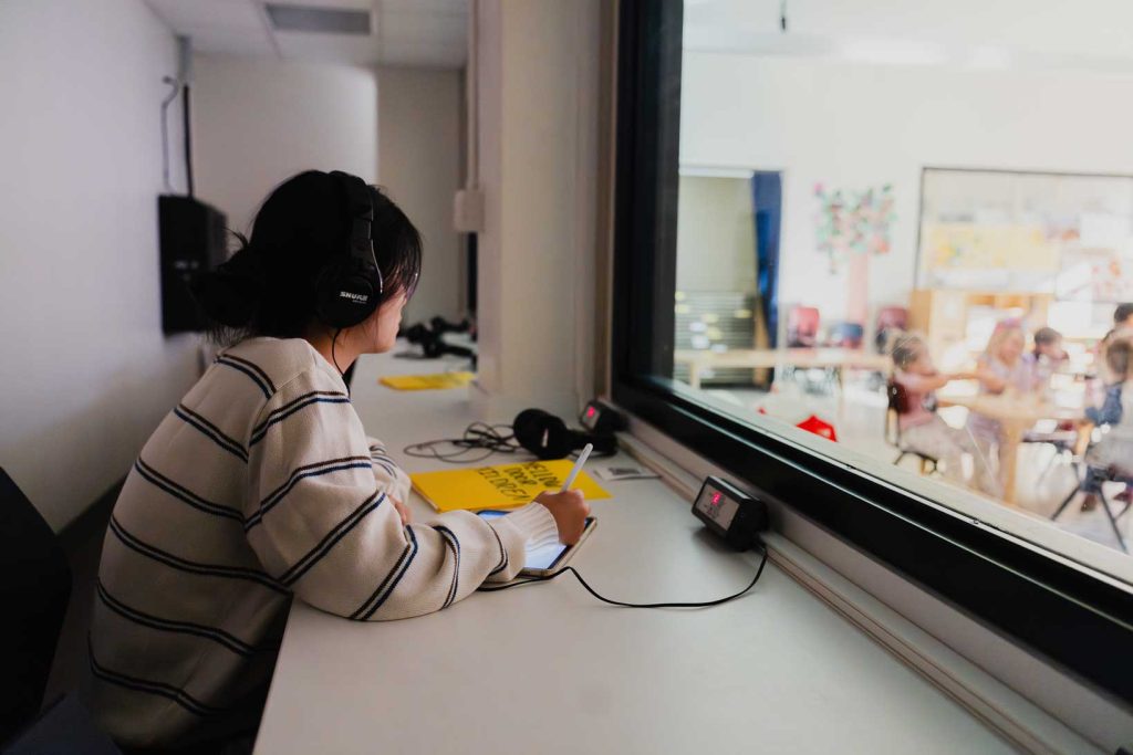 A woman with short brown hair wears a striped sweater and sits in an observation booth listening to the classroom through headphones.