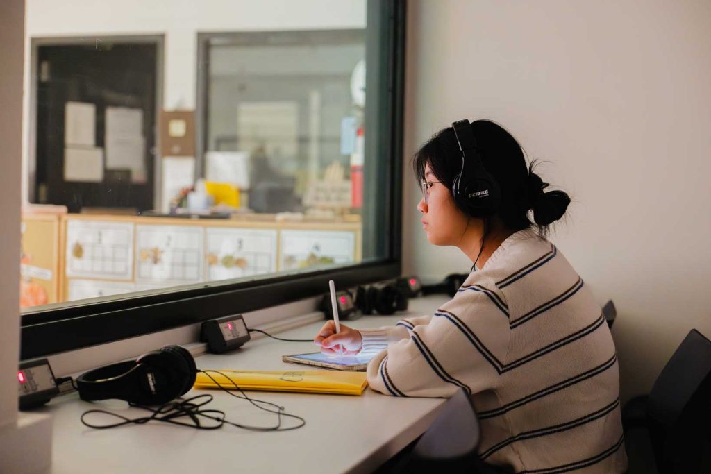 A woman with short brown hair wearing a striped sweater sits in an observation booth and records notes with a pencil. She is listening to the classroom through headphones.