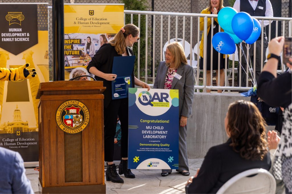 An early childhood professional receives an award recognizing the Child Development Lab for demonstrating quality through the Quality Assurance Report program at an outdoor ceremony.