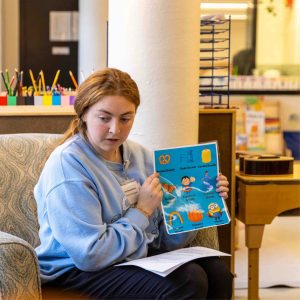 A woman with red hair sits in a chair and reads a book at the Child Development Lab.