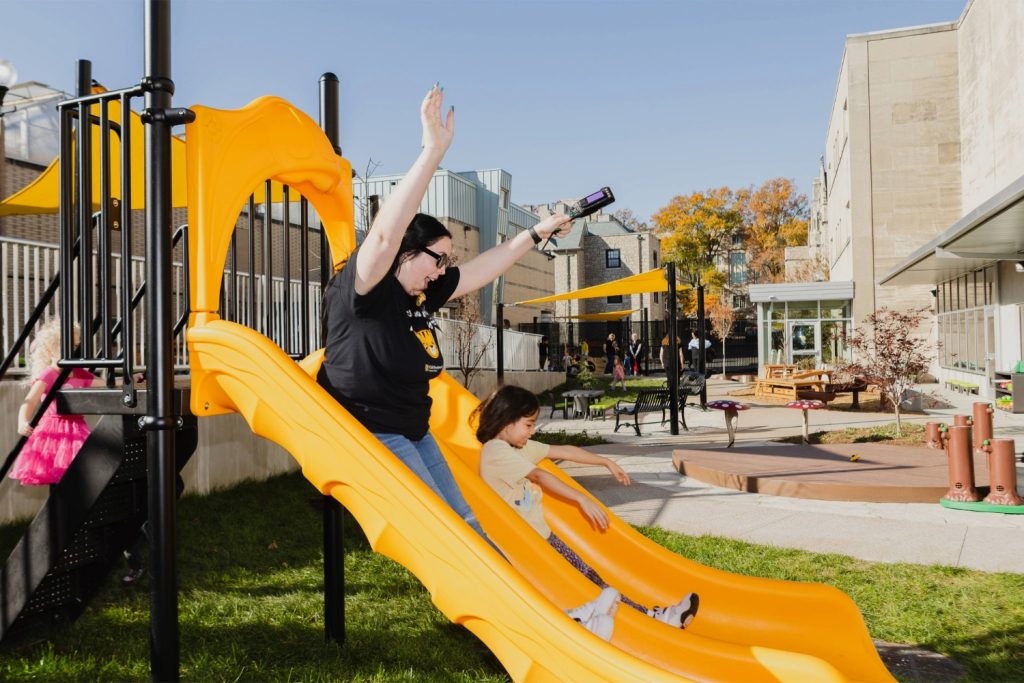 An early childhood teacher goes down the yellow slide next to a young child.