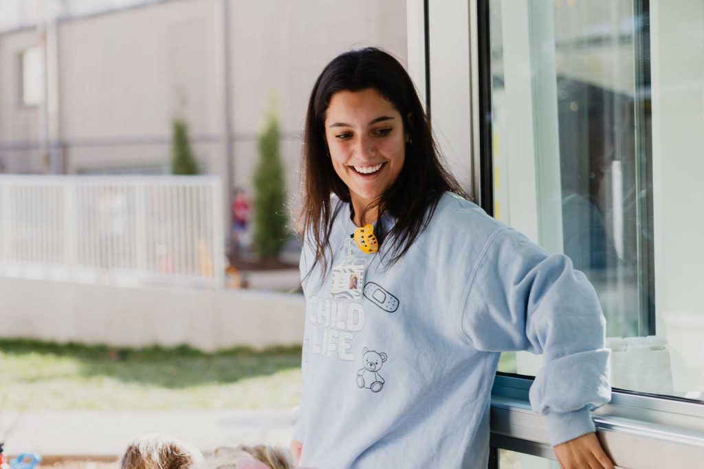 A woman with brown hair smiles while working outside at the Child Development Lab. She wears a yellow shirt under a grey sweatshirt.