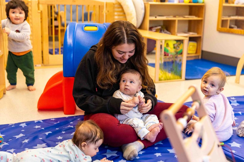 An early childhood teacher holds a baby while sitting on the ground with other babies and toddlers.