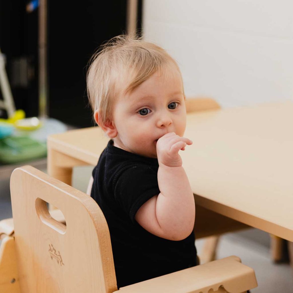 A baby sits in a chair at a table while sucking on their finger.