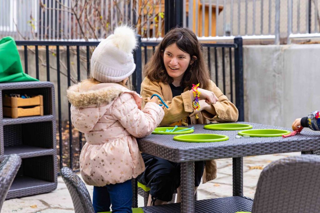 A child in a coat and hat works at a table with an early childhood teacher in the outdoor classroom at the Child Development Lab.