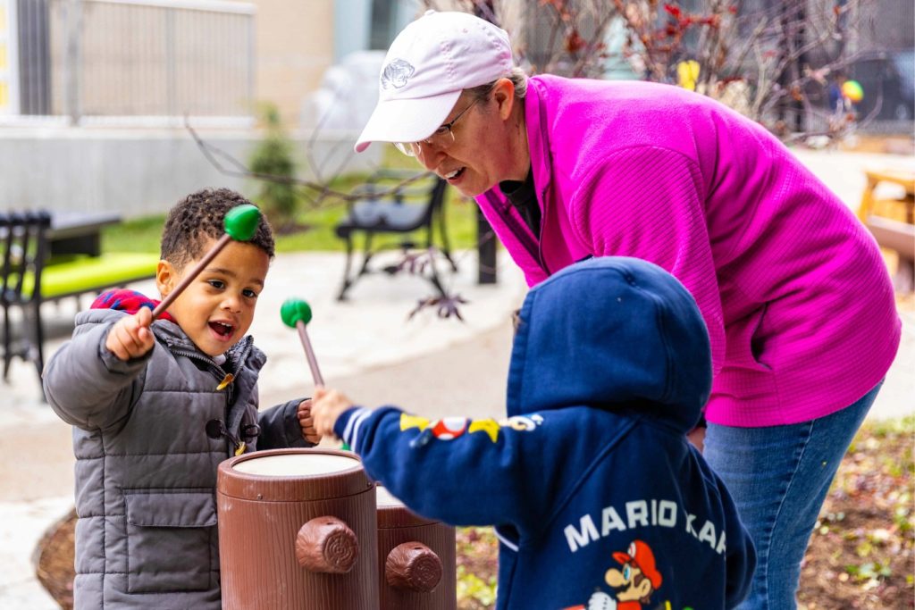 A woman in a pink coat and white hat kneels down to interact with two young children playing music in the outdoor classroom at the Child Development Lab.