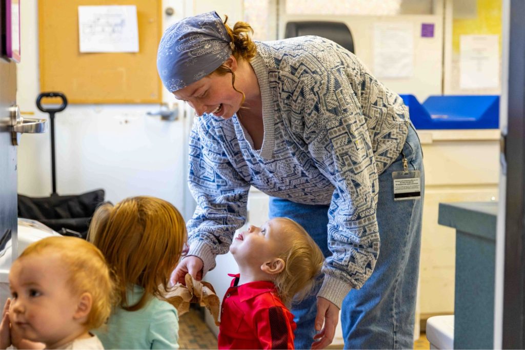 A woman with her hair in a bandana looks down while interacting with toddlers in the blue door classroom at the Child Development Lab.