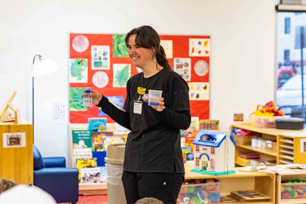 An early childhood professional smiles while holding two containers with a blue liquid for an activity in red door at the Child Development Lab.